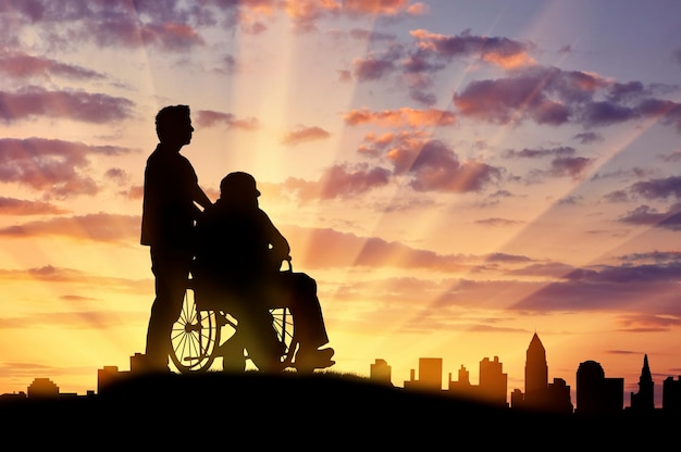 Concept of disability and old age. Silhouette of a man looking after a disabled person on a background of city sunset
