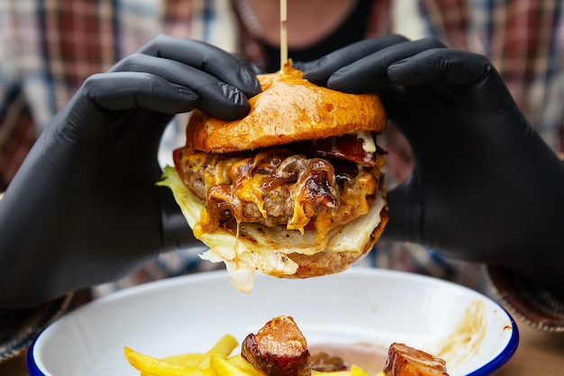 The concept of fast food and takeaway food A man in black latex gloves holds a juicy hamburger in his hands lies near french fries on a metal plate along with cheese sauce