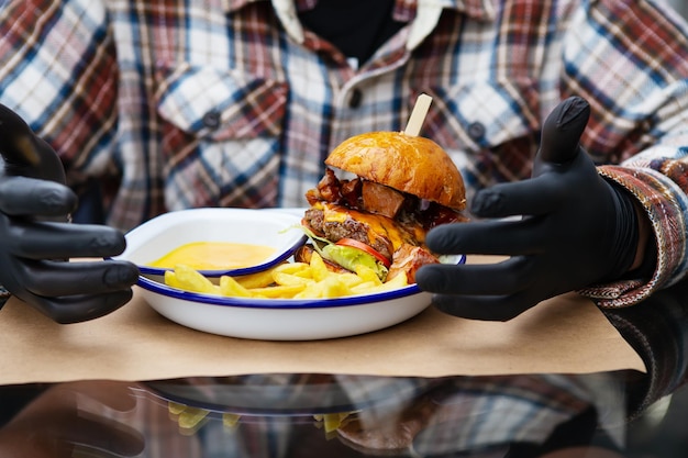 The concept of fast food and takeaway food a man in black latex\
gloves is ready to eat a juicy hamburger that lies near the french\
fries on a metal plate along with cheese sauce