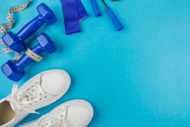 Photo the concept of fitness sport top view photo of white sports shoes elastic bands and blue dumbbells on isolated pastel blue background with empty space
