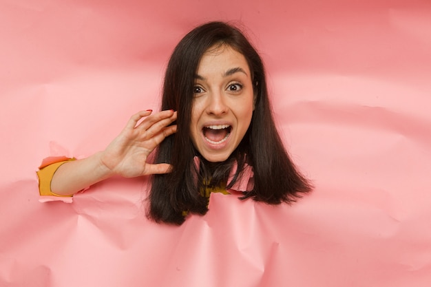Concept foto van jonge vrouw die papier scheurt en uit het gat gluurt, schreeuwend over commercieel aanbod op kopieerruimte, geïsoleerde roze kleurachtergrond.