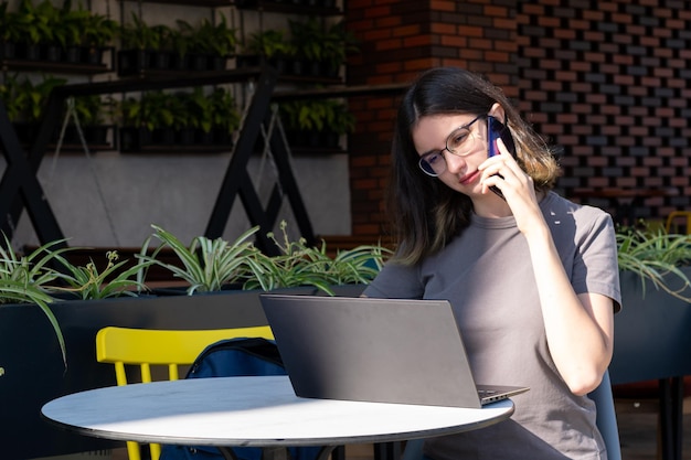 The concept of a freelancer A beautiful brunette student with glasses is talking on the phone using a laptop in a cafe