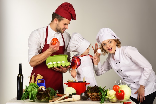 Concept of friendly family, mutual assistance, support, cooking - happy family in kitchen. Healthy food at home. Adorable kid in chef hat with her parents. Mother and father teaching boy how to cook.