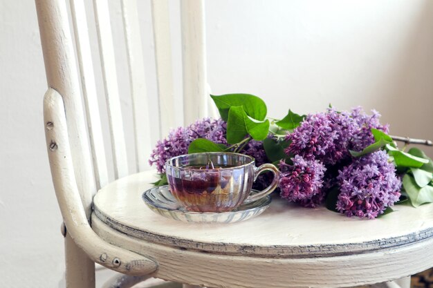 良い春の朝のコンセプト白い木製の椅子の側面図にライラックの花束とお茶のカップ