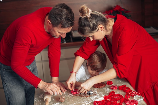 Concept, groepswerk. Een jong gezin, vader, moeder en kind verspreidden de gesneden zelfgemaakte deegkoekjes op een bakplaat. Vrolijke kerstmomenten gekleed in het rood. Jeugd. Zoon van 4 jaar.