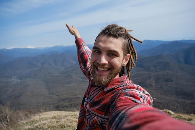 Concept of healthy active lifestyle Young travel blogger with dreadlocks went hiking in mountains Man enjoys views of nature takes video or selfie photo on phone or camera Wideangle view portrait