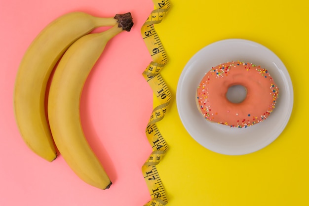 Concept of healthy or fast eating. Bananas, tasty colorful doughnut and tape measure on bright yellow and pink background. Top view, minimal style