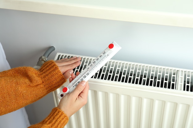 Concept of heating season with girl holds thermometer near radiator.