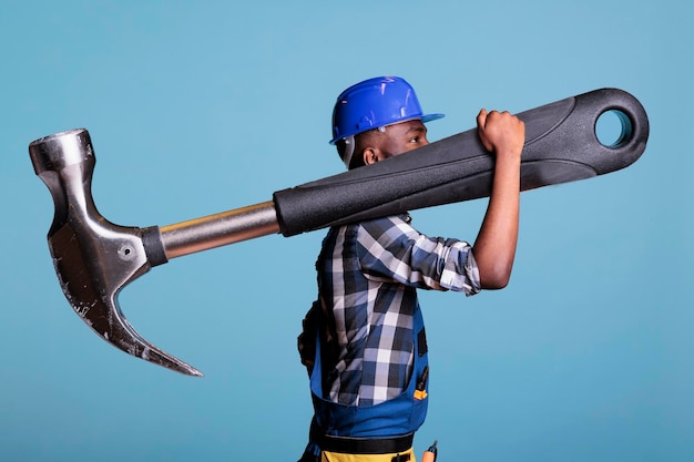 Concept of heavy work, African american builder with giant hammer in studio shot. Strong man heavy work Construction worker carrying huge work tool wearing coveralls and hard hat, construction industr