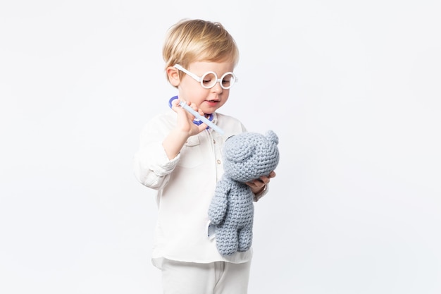 Photo concept of kid medicine, health. child doctor with glasses, stethoscope gives the bear medicine on white background with copy space.