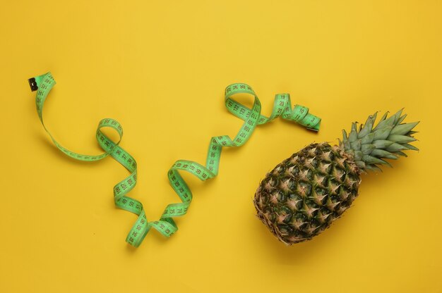 The concept of losing weight. Pineapple wrapped with measuring tape on yellow background. Healthy eating. Top view