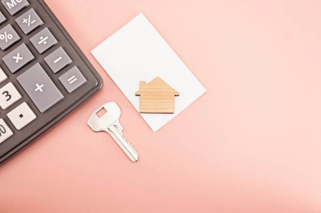 The concept of mortgage, sale and rental of housing and real estate. Mortgage credit lending. Calculator with a wooden house and a key on a delicate pink background. Copy space. Mock-up.