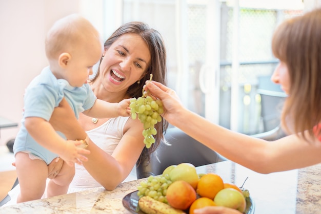 The concept of motherhood, nanny, infancy and childhood. Indoor shot in the kitchen. Two women and a child in their arms, the child is offered fruit, happy family