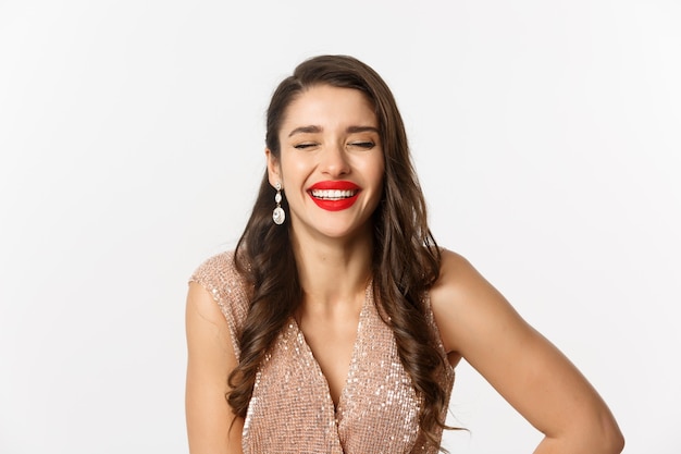 Concept of New Year celebration and winter holidays. Close-up of elegant woman in dress, with red lips, laughing and looking happy, standing over white background.