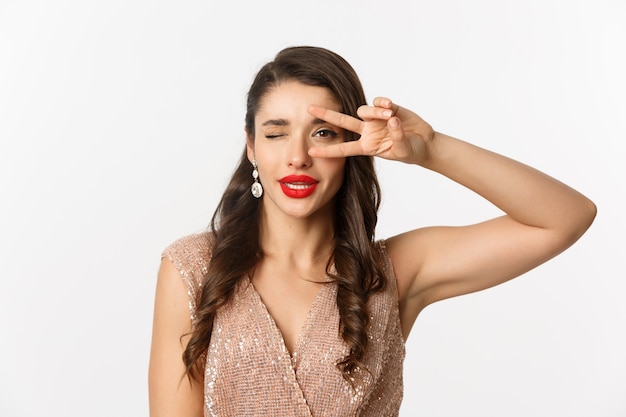 Concept of New Year celebration and winter holidays. Close-up of glamour woman in party dress, red lips, showing peace sign and winking sexy at camera, white background.