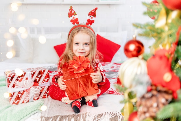 The concept of the new year and Christmas, a girl child on the bed at home with gifts waiting for the new year or Christmas in a red sweater and smiling with happiness