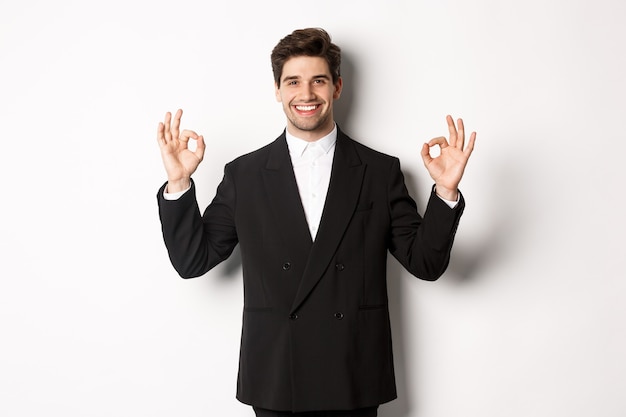 Concept of new year party, celebration and lifestyle. Portrait of handsome attractive man in black suit, smiling and showing okay signs, approve and recommend something, white background