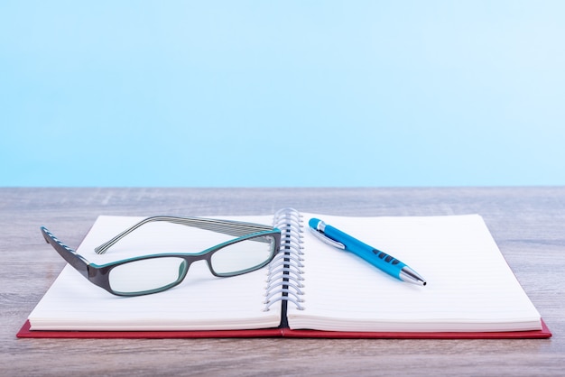 Concept onderwijs of bedrijf: Open dagboek of notitieboek met moderne bril en blauwe pen op de houten tafel geïsoleerd op blauwe achtergrond.