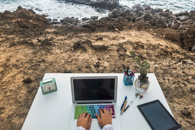 Concept of outdoor alternative office workplace Digital traveler nomad people lifestyle Above view of desk with laptop computer and hands writing on with ocean view New normal small business