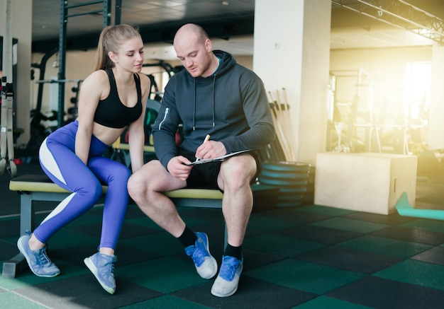 The concept of personal training. Instructor notes in notebook results  training sporty blonde woman sitting on a bench. Smiling sports couple