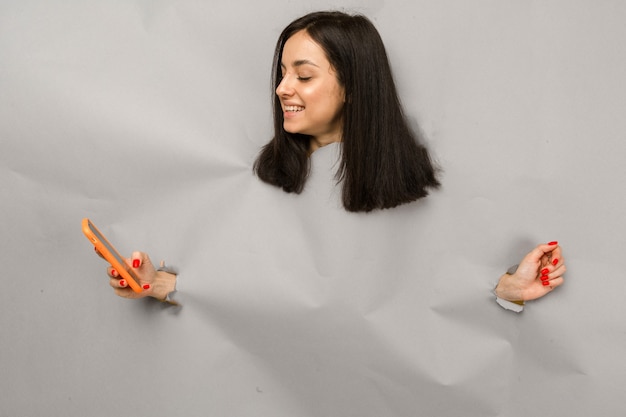 Concept photo of young female tearing paper and peeking out hole, holding smartphone, isolated grey color background.