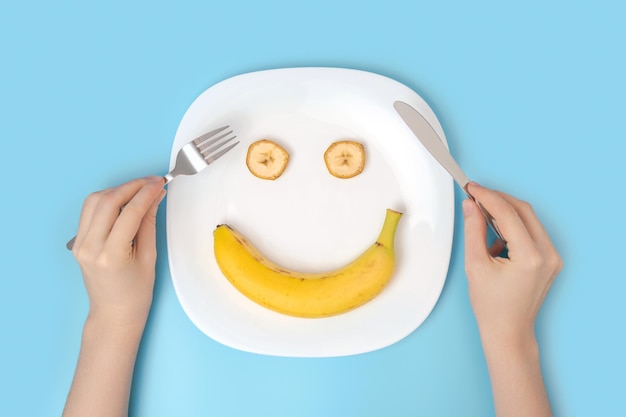 Foto il concetto di un atteggiamento positivo nei confronti della dieta e del mangiare sano le mani delle donne con le posate su un piatto con una banana su sfondo blu vista dall'alto