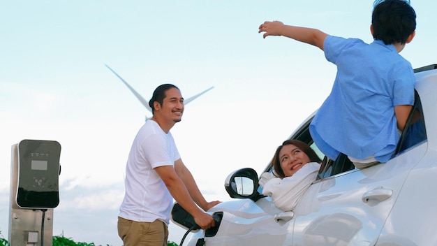 Concept of progressive happy family enjoying their time at wind farm with electric vehicle Electric vehicle driven by clean renewable energy from wind turbine generator for charging station