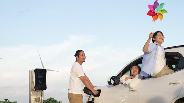 Concept of progressive happy family enjoying their time at wind farm with electric vehicle Electric vehicle driven by clean renewable energy from wind turbine generator for charging station