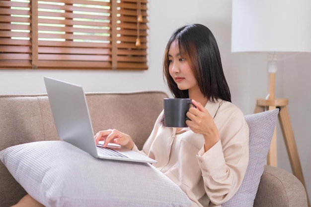 Концепция отдыха дома Молодая азиатка просматривает социальные сети на ноутбуке и пьет кофе
