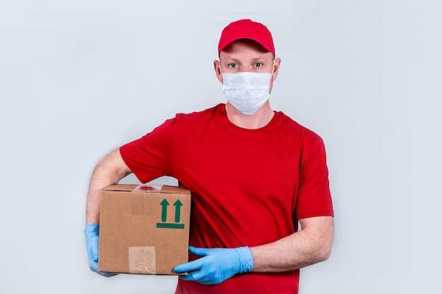 Il concetto di consegna sicura. un corriere in uniforme rossa e maschera protettiva e guanti tiene una scatola di cartone, consegna senza contatto degli ordini