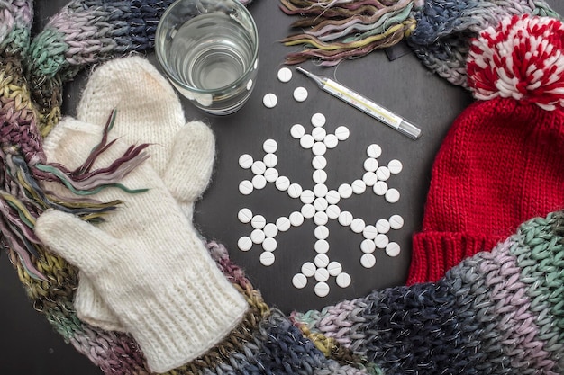 Foto fiocchi di neve concettuali realizzati con compresse medicinali, vestiti invernali colorati vicini, un bicchiere d'acqua e un termometro.