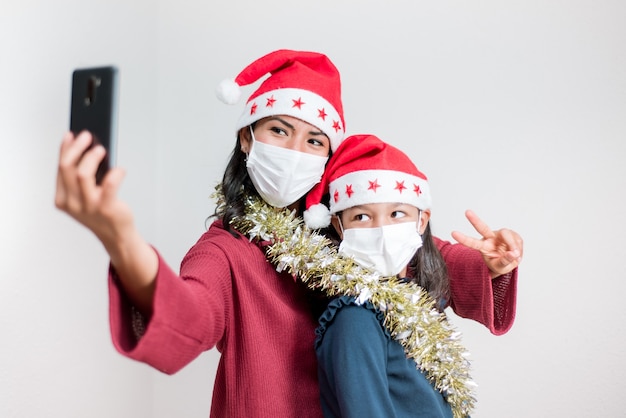 크리스마스 코로나 바이러스 동안 사회적 거리의 개념. 라틴어 어머니와 딸이 얼굴 마스크를 쓰고 휴대폰으로 셀카를 찍습니다.
