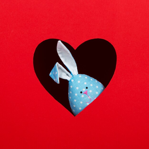 The concept of spring, happy Easter, eared blue rabbit handmade, peek out of a hole in the shape of a heart on a red background. Copy space.