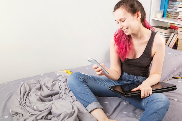 Concept - thuiswerken, zelfisolatie. het jonge meisje zit in bed onderzoekend telefoon en glimlacht, naast laptop