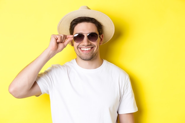 観光と休暇の概念。幸せそうに見える、サングラスと夏の帽子をかぶって、黄色の背景の上に立っているハンサムな男の観光客のクローズアップ。