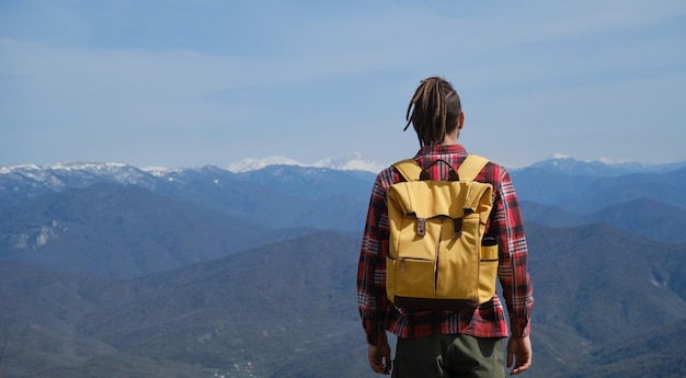 Concept of travel and healthy active lifestyle Man with yellow backpack stands on top of hill and enjoys views of nature Caucasus Rear view Young guy with dreadlocks went hiking in mountains