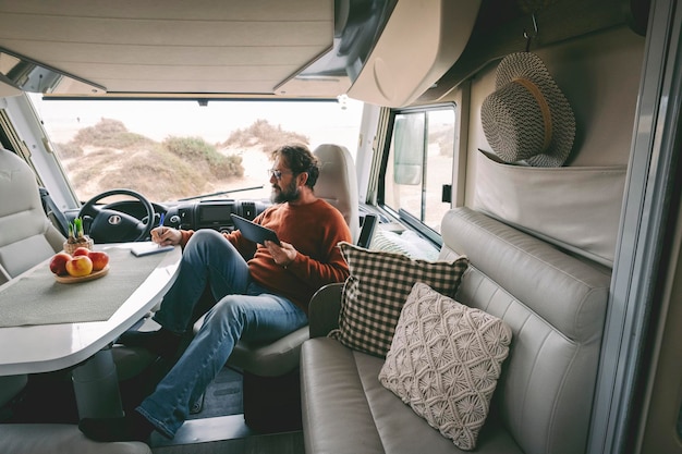 Concept of travel lifestyle modern people and smart working\
digital nomad job activity handsome man use laptop computer sitting\
and relaxing inside a camper van vehicle parked at the beach\
freedom