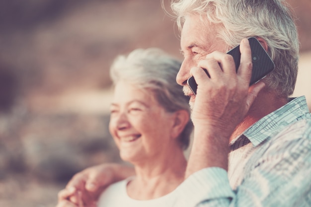 concept of vacation, technology, tourism, travel and people - happy senior couple with cellular phone on phone on pebble beach laughing and joking. White hair