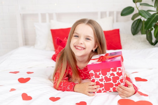 the concept of Valentine's day a cute child girl is sitting on the bed at home in red pajamas and holding a gift in her hands and smiling or laughing with happiness congratulating on the holiday