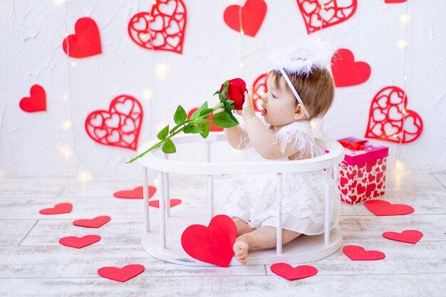 Il concetto di san valentino una gioiosa bambina di sei mesi è come un angelo tra cuori rossi e con una rosa tra le mani sorridente