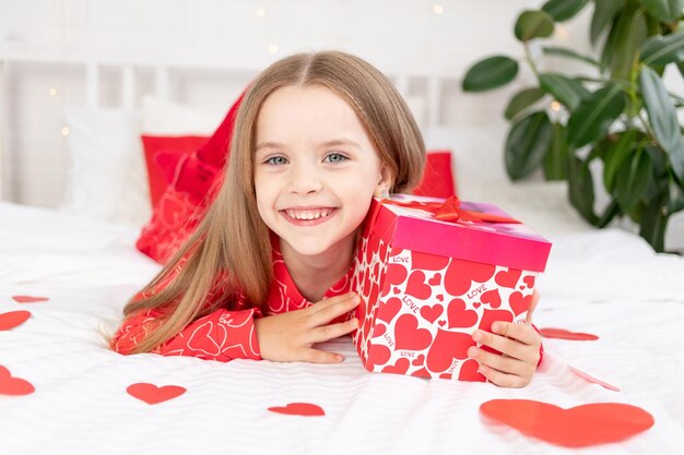 The concept of Valentines day a cute child girl is sitting on the bed at home in red pajamas and holding a gift in her hands and smiling or laughing with happiness congratulating on the holiday