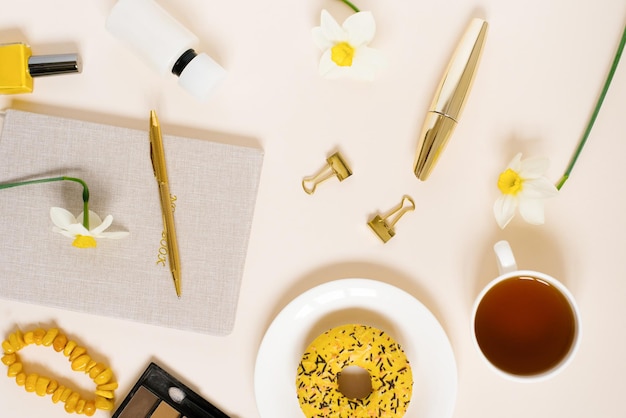 Concept van een lenteblog Een kopje thee een donut narcis bloemen briefpapier cosmetica op een beige achtergrond Plat liggende vrouwelijke tafel met bovenaanzicht