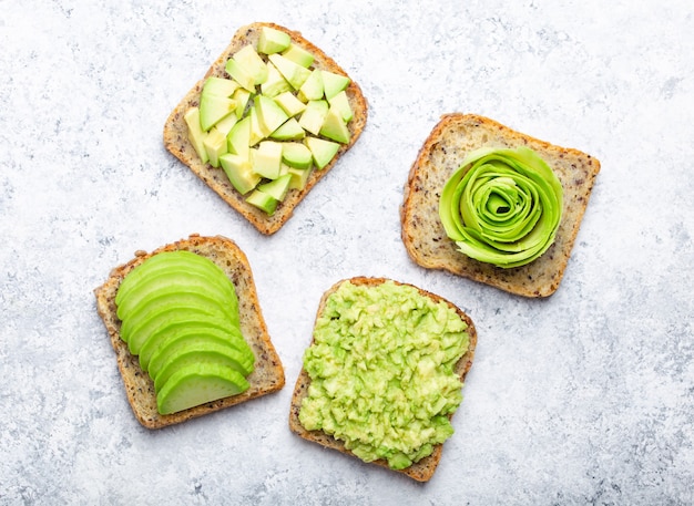 Concept van gezonde ontbijttoosts met ingeslagen avocado, plakjes, brokken en roos. Verschillende manieren om heerlijke avocado-sandwich te maken en te serveren, bovenaanzicht, witte stenen achtergrond, close-up