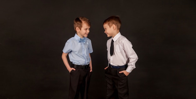 Concept van onderwijs, opvoeding, mannelijke vriendschap en broederschap. Twee kleine stijlvolle jongens broers en vrienden, sprekend en gelukkig samen, op zwarte achtergrond. Portret van 2 jongens op weg naar hetzelfde doel