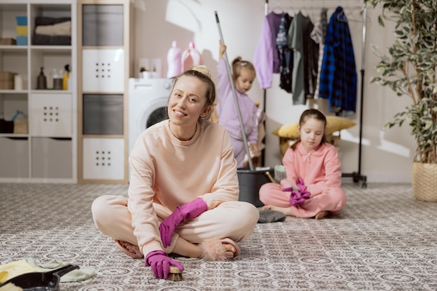 Concept van thuis schoonmaken met kind Moeder zit op de badkamervloer in de wasruimte en zij en dochters schrobben de vloer met een borstel en vloeibaar wasmiddel Kinderen leren schoonmaken