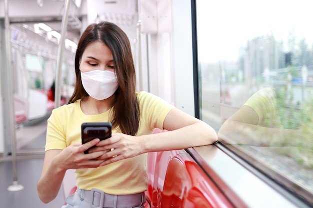 Concept van verkeer en vervoer aziatische vrouwelijke passagier die een masker draagt met een smartphone die online via internet chat ze zit in de skytrain die in de stad pendelt