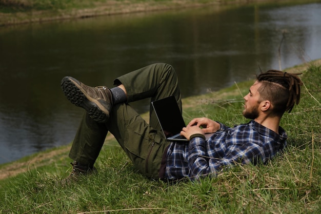 Concept van werken op afstand en zaken met behulp van internettechnologieën jonge blanke man met dreadlocks en baard liggend op de rug bij het opruimen op de rivieroever in het voorjaarspark en werkend op laptop