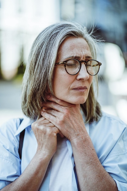 Concept van ziekte door een volwassen dame die de keel vasthoudt Vrouw lijkt ziek en moe met een vermoeide uitdrukking op het gezicht Koud getinte kleurenpalet draagt bij aan het algemene gevoel van ziekte en ongemak