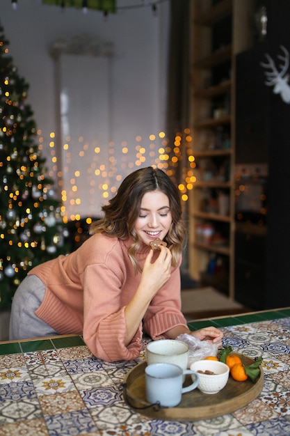 Concept voor kerstfotografie, gelukkige jonge vrouw in warme trui eten in donkere keuken