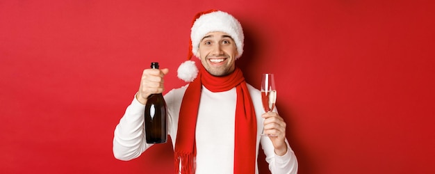 Concetto di vacanze invernali, natale e stile di vita. primo piano di un bell'uomo allegro, che tiene in mano una bottiglia di champagne e un bicchiere, fa un brindisi per il nuovo anno e festeggia, sfondo rosso.